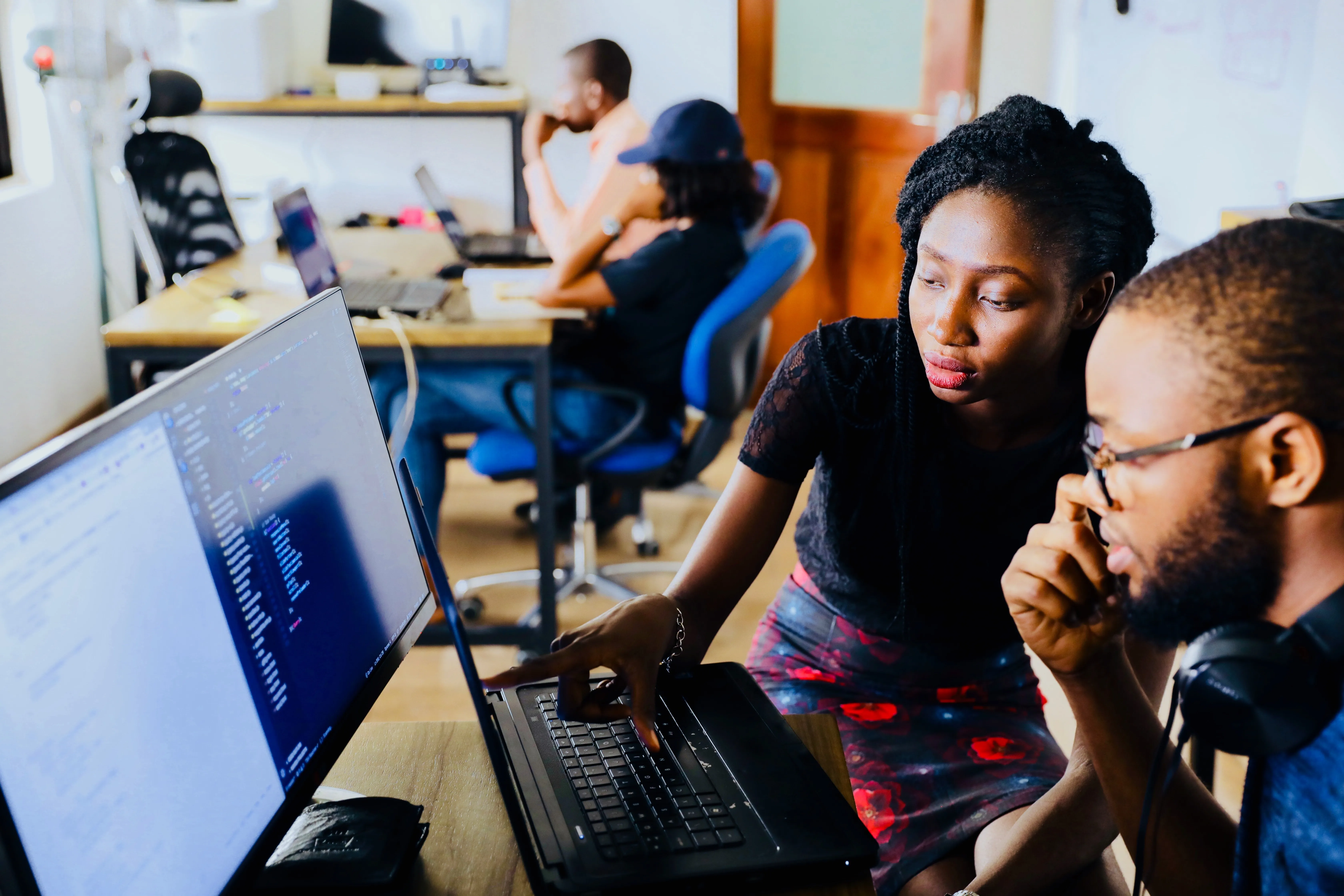 A woman is sitting beside a man in glasses at a computer. She is pointing to something on the computer monitor that we cannot read.
